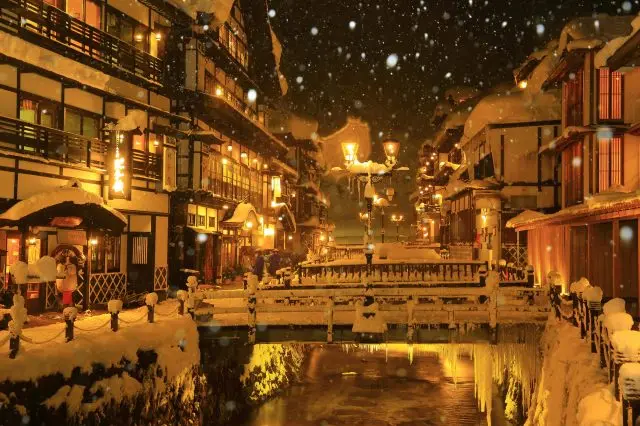 The snow-covered hot spring town glows softly with lights, creating a dreamy scene