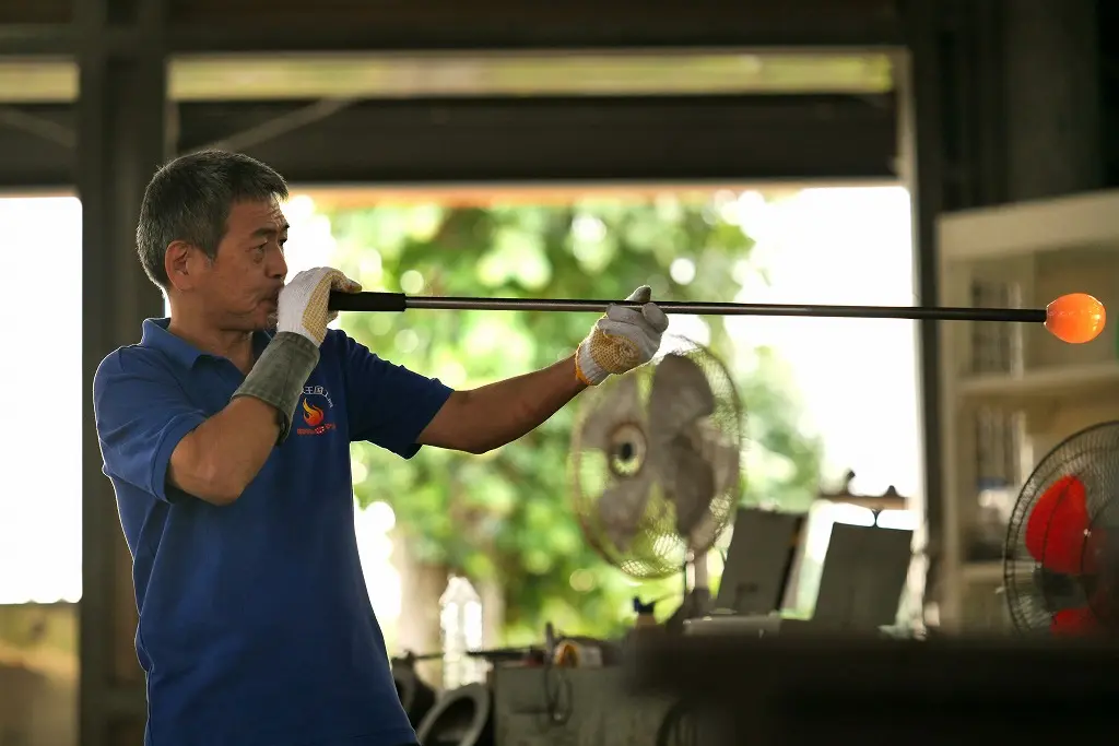 ガラス工房で職人が竿を使い、溶けたガラスを吹き上げて成形する作業の様子