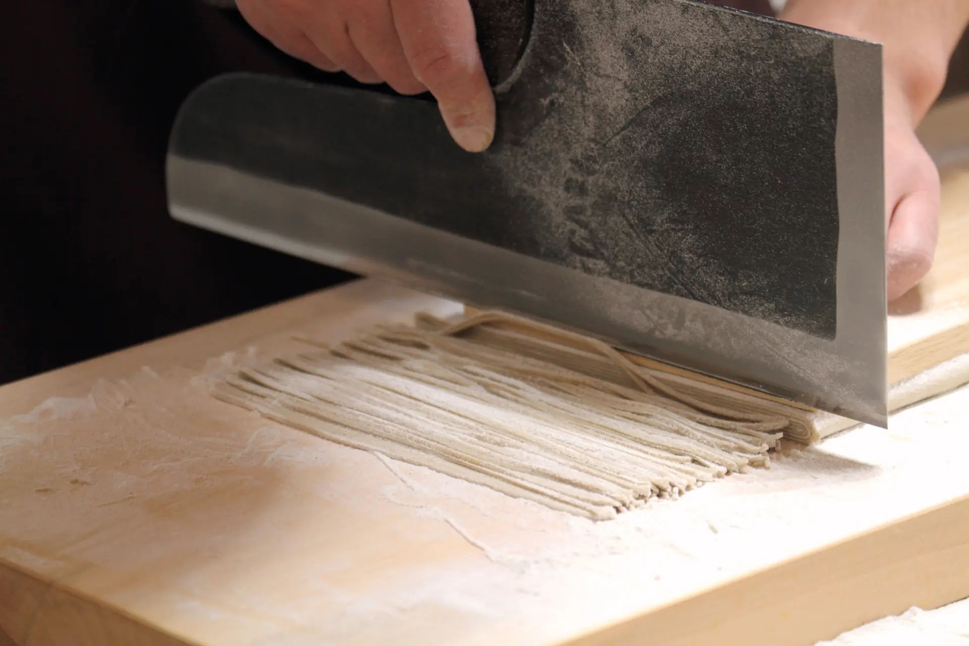 打ちたてのそば生地を包丁で細く切り分ける作業。粉をまとった麺がまな板の上に整う