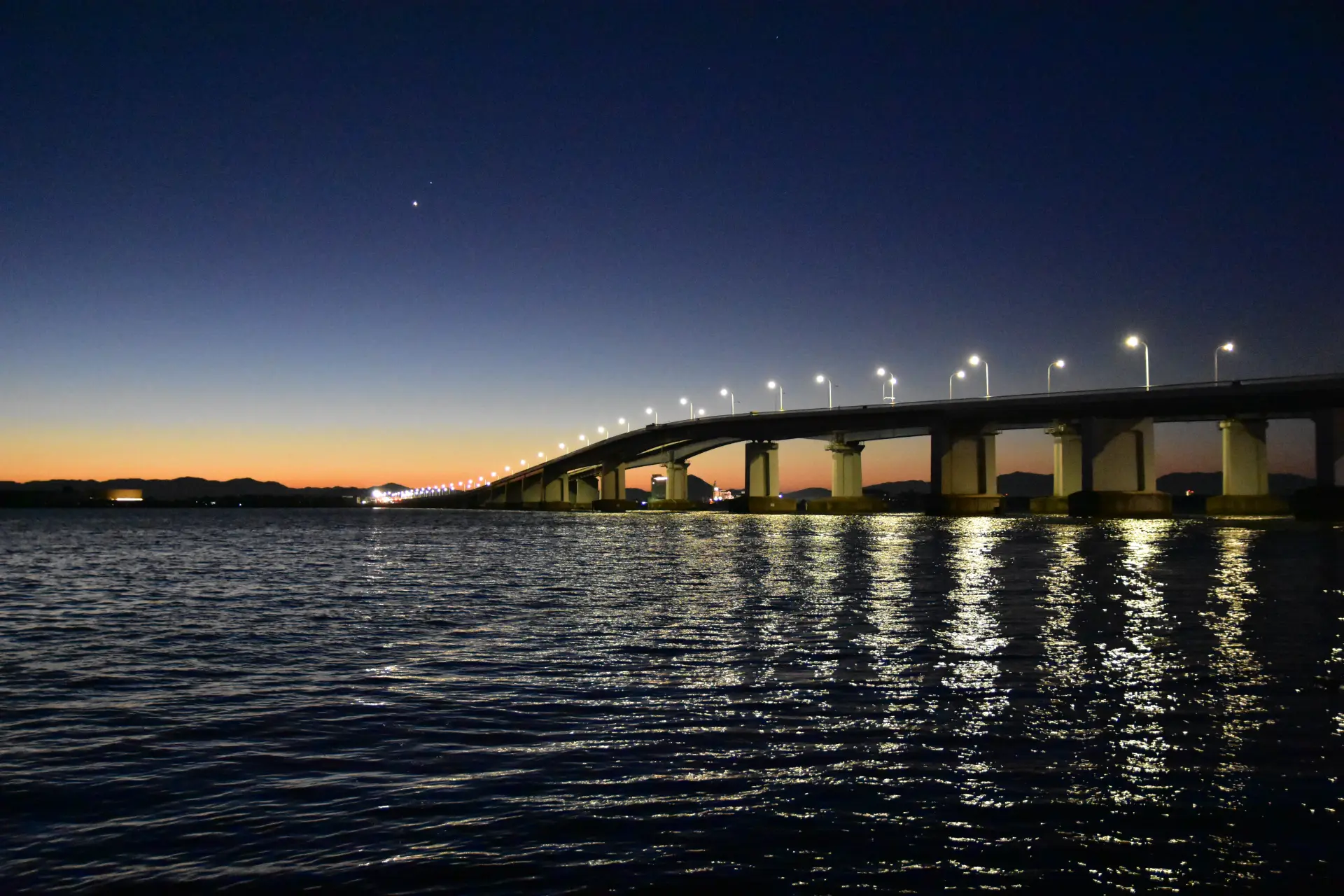 橋のライトと水面に映る光が重なり、静かな湖上に広がる夜の景観を描く