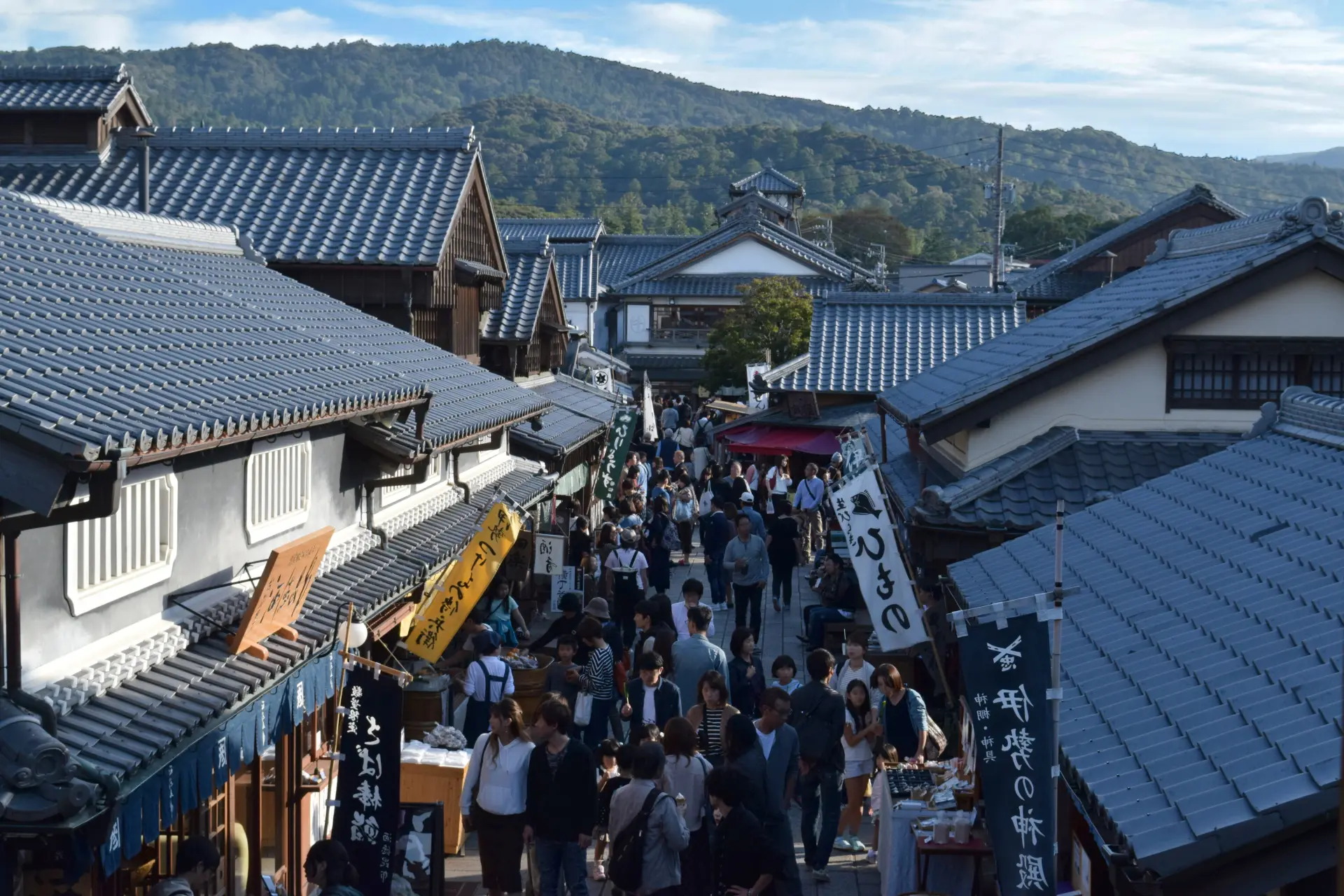 瓦屋根の町並みに店舗が並ぶ通り。人の行き交いと食べ歩きのにぎわい